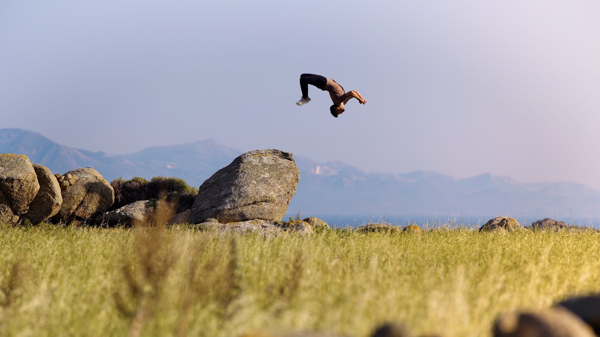 A figure mid-flip over a golden grass field with mountains behind — a frame from Sage Moore's cinematography.